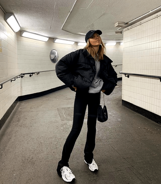 chica rubia usando una gorra de beisbol, sudadera gris, chamarra negra, bolso pequeño, leggings negros y tenis deportivos blancos