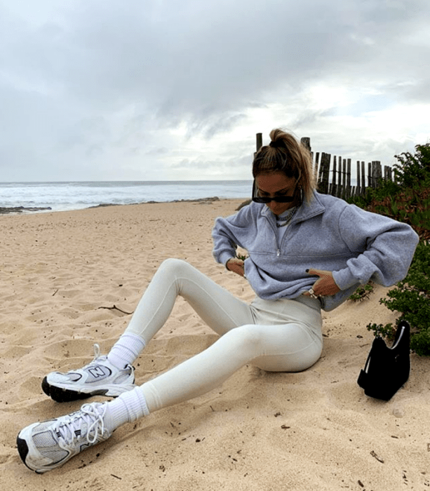 chica rubia usando lentes de sol, sudadera gris, leggings blancos, tenis deportivos blancos y minibolso negro