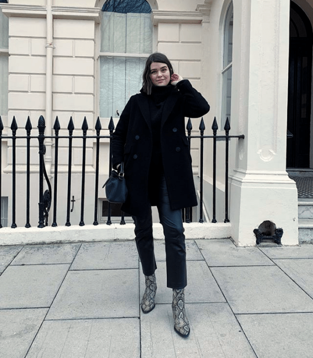 chica de cabello corto oscuro usando un abrigo negro, top negro de cuello alto y pantalones de mezclilla con botines de piel y bolso negro 