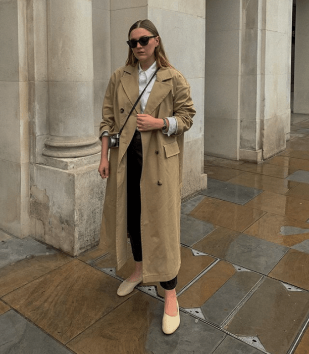 chica rubia usando un abrigo café claro con lentes de sol, pantalón negro, camiseta de cuello alto blanca y ballerinas beige