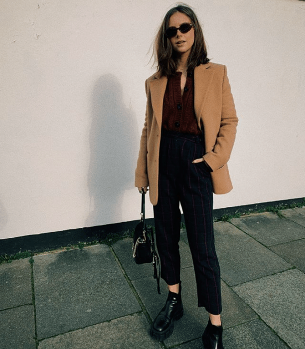 chica de cabello largo castaño usando un blazer de vestir beige, top guinda, pantalones de vestir, bolso pequeño de cuero y botines de cuero negros