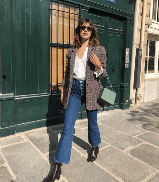 chica de cabello castaño largo usando lentes de sol, camisa de vestir blanca, chaqueta de vestir de cuadros, jeans acampanados y botines cafés y bolsa de mano verde