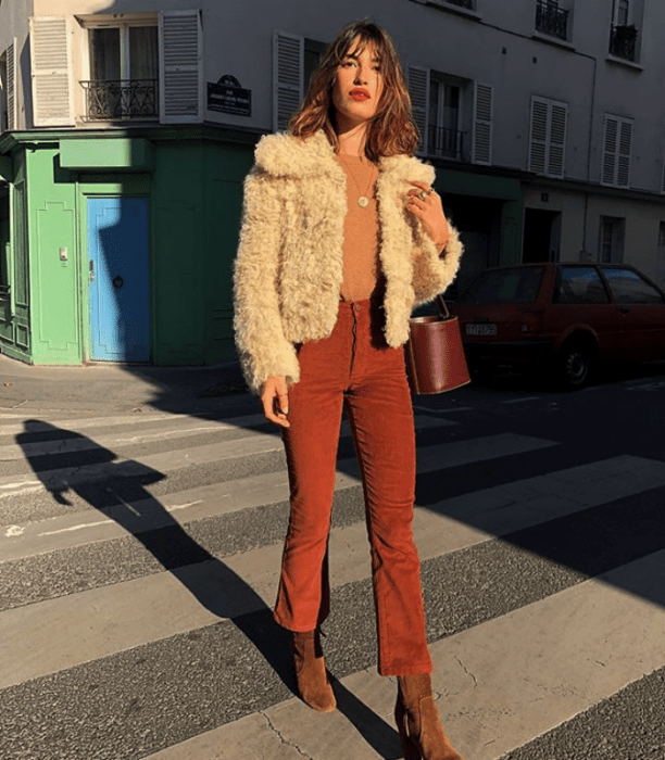 chica de cabello largo castaño usando un abrigo de peluche blanco con top anaranjado y pantalones color rojo y botines