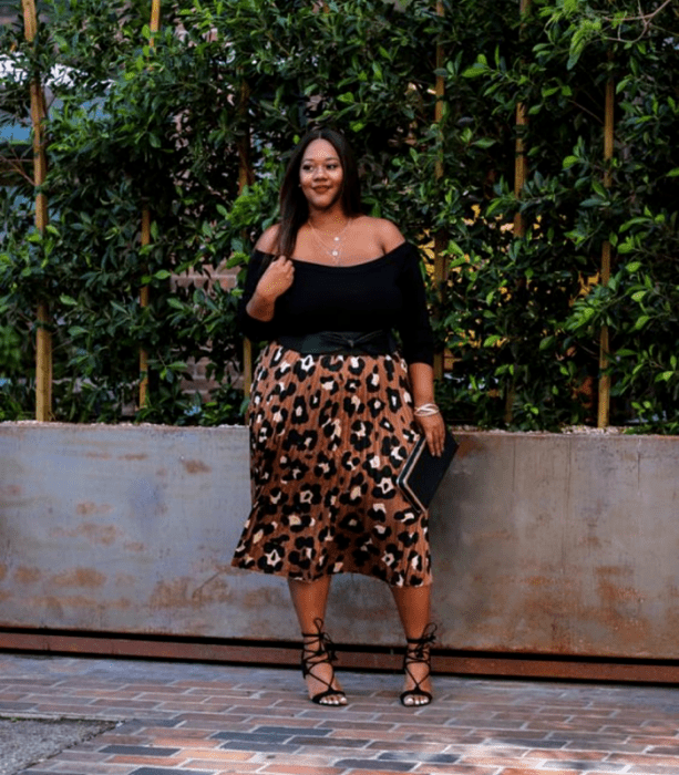 chica curvy morena de cabello largo oscuro usando una blusa sin hombros negra de 3/4 con falda midi de animal print, sandalias negras de tacón y bolso de mano negro