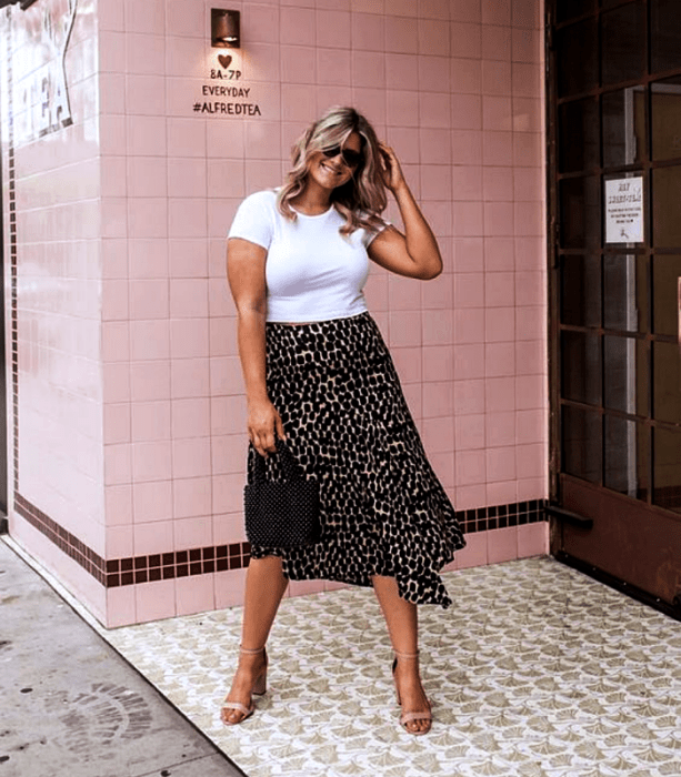 chica curvy usando lentes de sol, camiseta blanca de manga corta, falda midi de animal print, bolso de cuentas negro y sandalias de tacón beige