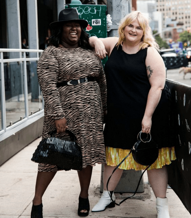 chicas curvy, una rubia con vestido negro largo sin mangas y falda amarilla con naranja debajo, botines blancos y bolso negro, otra chica morena con sombrero negro, vestido de animal print largo de manga larga, bolso negro de mano y sandalias de tacón negras