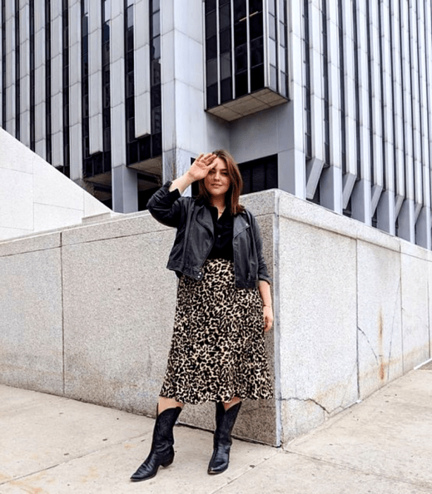 chica curvy de cabello castaño usando una camiseta negra, chamarra de cuero, falda larga de animal print y botines vaqueros negros 