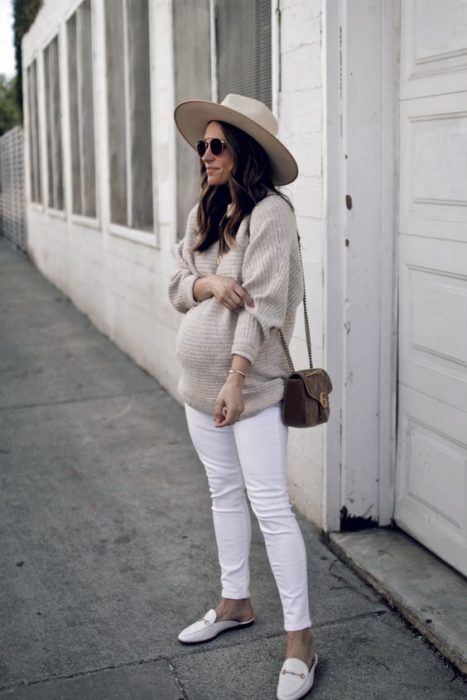 Chica usando un outfit de maternidad de jeans blancos, sueter color café y sombrero 