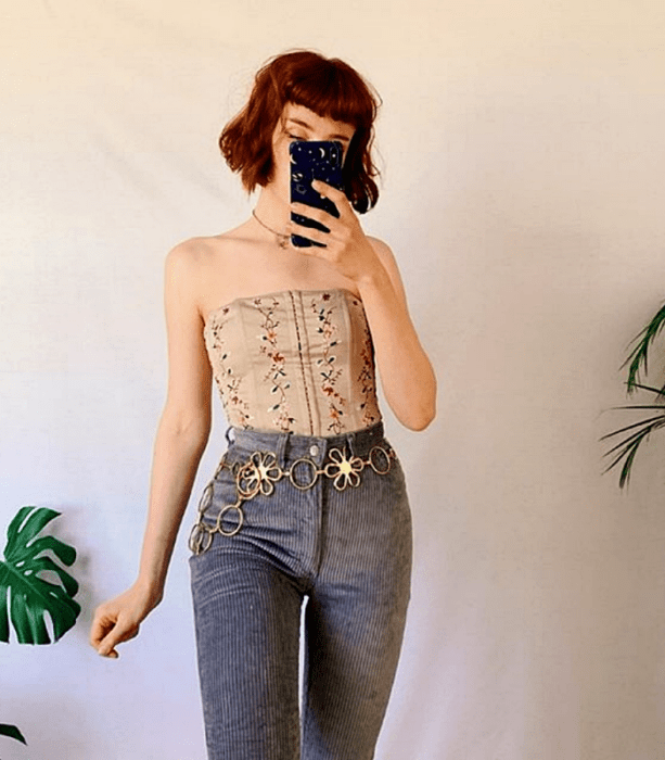 chica pelirroja usando un top beige tipo corset con jeans de pana grises y cinturón metálico con flores