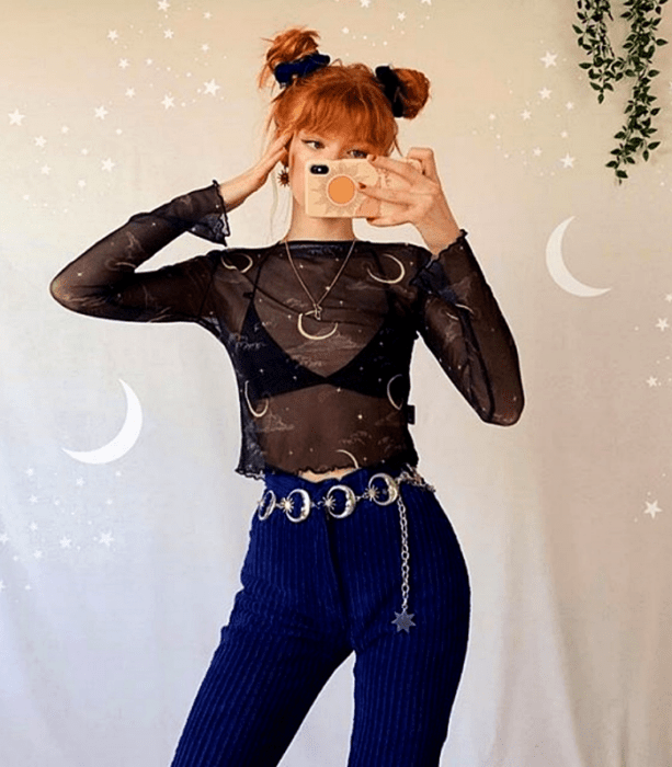 chica pelirroja con space buns, top de manga larga semitransparente y sostén negro, pantalones de pana azul marino y cinturón metálico con lunas