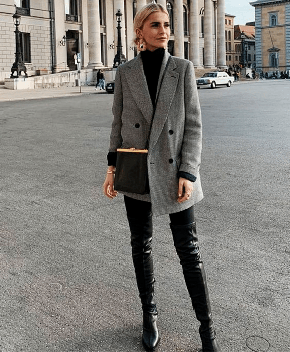 chica rubia usando top negro de cuello alto, blazer gris oversized, bolso negro, leggings negros, botas largas de cuero negras