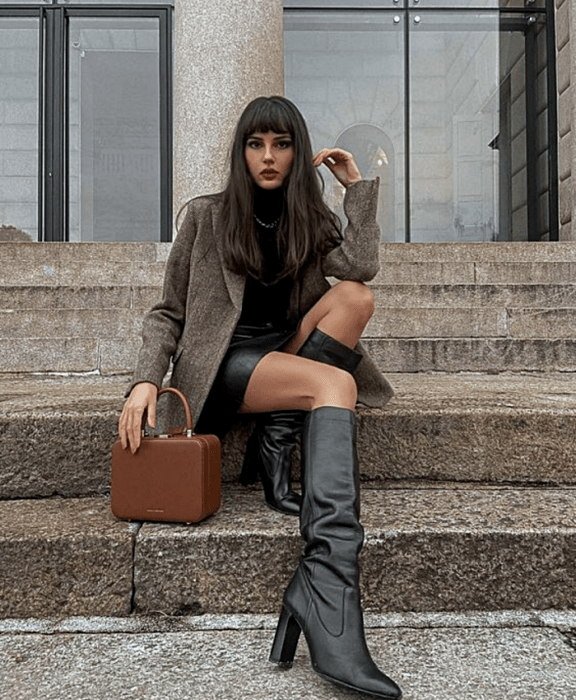 chica de cabello castaño largo usando un top negro de cuello alto, blazer café largo, mini falda de cuero, botas largas de cuero con tacón y bolso café de mano