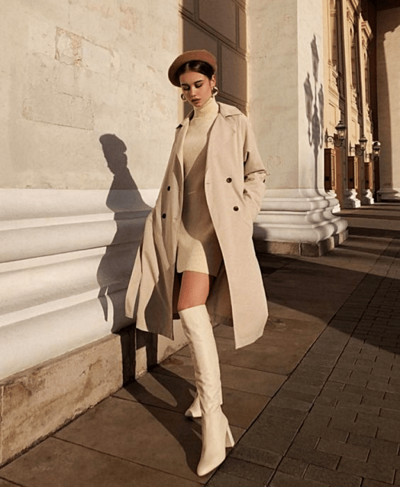 chica de cabello castaño usando un beret café, vestido de suéter de cuello alto beige, abrigo beige largo, botas blancas de cuero blanco con tacón