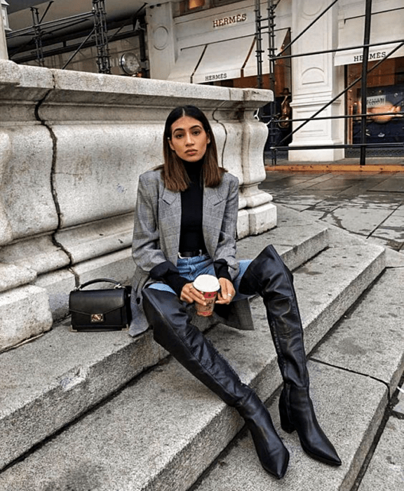 chica de cabello castaño usando un top negro de cuello alto, blazer gris saco oversized, jeans a la cintura, botas largas de cuero con tacón, y bolso negro