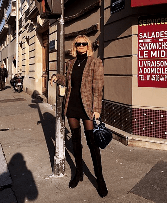 chica rubia usando lentes de sol, vestido negro de cuello alto, abrigo oversized café con cuadros, medias negras, botas largas negras de cuero con tacón y bolso de mano de terciopelo