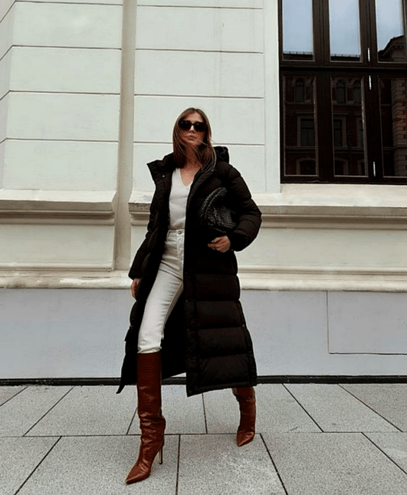 chica de cabello castaño usando lentes de sol, top blanco de cuello en V, abrigo chamarra negra larga, jeans blancos, botas largas cafés de cuero con tacón, y bolso negro de mano