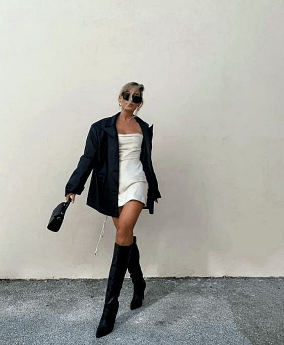 chica rubia usando lentes de sol, vestido mini blanco, abrigo de cuero negro, botas largas de cuero con tacón y bolso mini negro