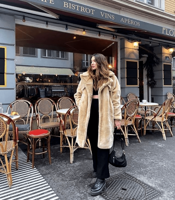 chica de cabello largo claro con abrigo teddy largo beige, crop top negro, pantalón de vestir negro y botas de cuero de suela gruesa y mini bolso de mano negro