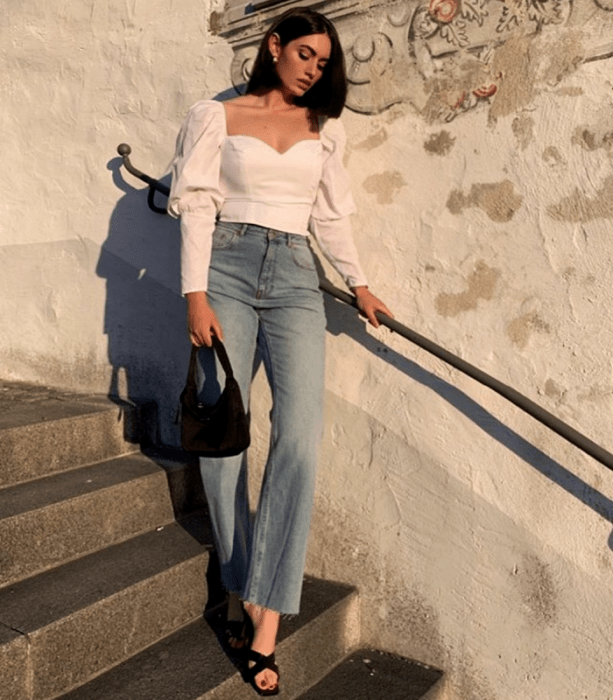 chica de cabello castaño usando una blusa blanca de manga larga, jeans a la cintura, sandalias de tacón y bolso mini negro