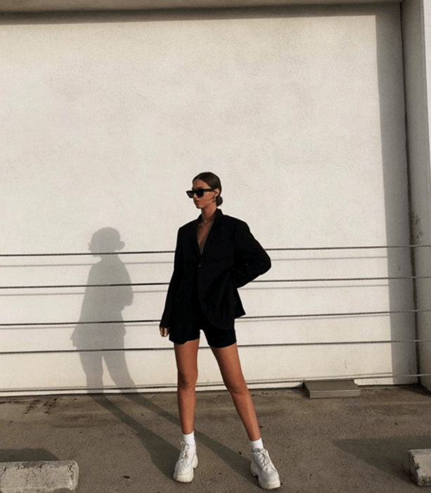 chica de cabello castaño usando lentes de sol, blazer negro oversized, shorts de ciclismo negros y tenis deportivos blancos de suela gruesa