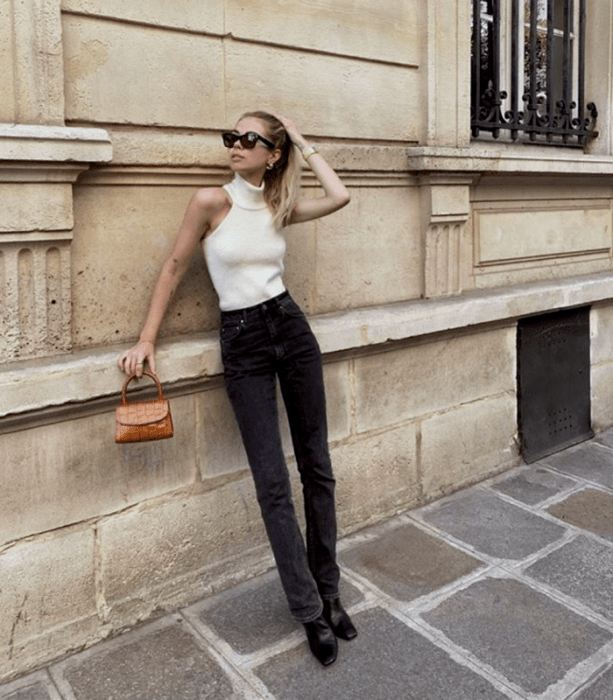 chica rubia con lentes de sol, top tejido blanco de cuello alto sin mangas, jeans a la cintura negros, botines negros y mini bolso naranja