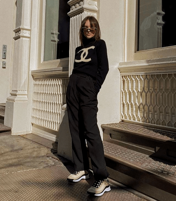 chica con cabello castaño usando lentes de sol, suéter negro con logo de chanel, pantalones negros de vestir, tenis deportivos beige con negro y suela blanca