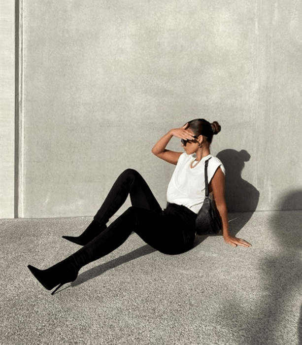 chica de cabello castaño usando lentes de sol, top blanco sin mangas, leggings a la cintura negros, botines de tacón negros y bolso negro de cuero