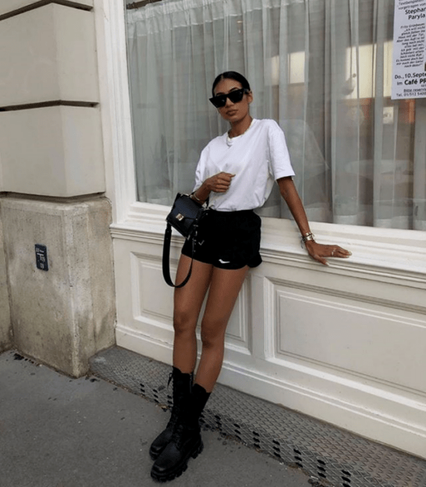 chica morena de cabello castaño usando lentes de sol, camiseta blanca, shorts negros cortos, bolso mini de cuero, botines largos de suela gruesa 