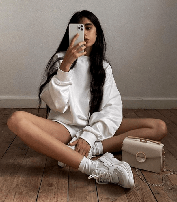 chica de cabello castaño largo usando sudadera blanca, shorts blancos, tenis deportivos blancos y bolso beige blanco tomándose una selfie