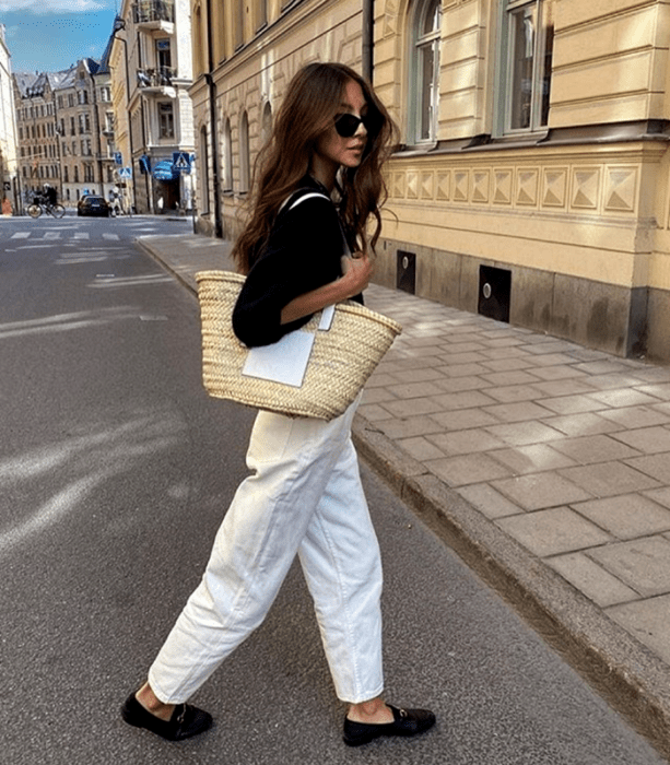 chica de cabello castaño largo con lentes de sol, suéter negro, bolso tejido de rafia grande, pantalones de mezclilla blancos holgados y zapatos tipo loafers negros