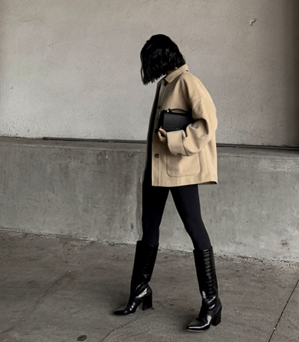 chica de cabello oscuro corto usando una chaqueta oversized beige, bolso mini de  mano negro, leggings negros y botas largas de tacón de piel