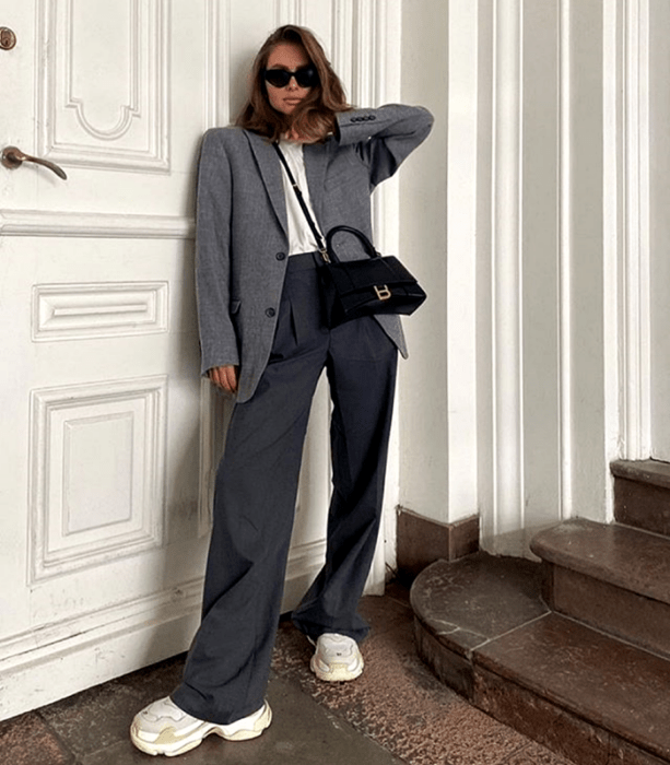 chica de cabello claro usando lentes de sol, top blanco, blazer oversized gris, pantalones grises de vestir, tenis blancos deportivos de plataforma gruesa y bolso mini negro 