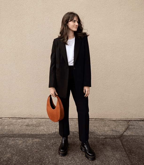 chica de cabello castaño usando una camiseta blanca, blazer negro, pantalones de vestir negros, botas de suela gruesa negras y bolso de mano naranja