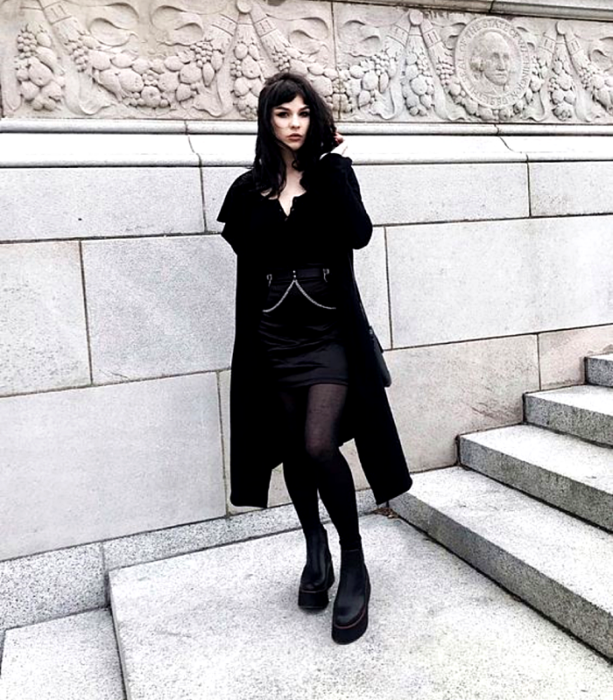 chica de cabello negro usando un top negro, abrigo negro, minifalda negra con cinturón negro con cadena, medias negras y botines negros de piso