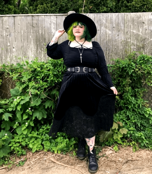 chica curvy de cabello verde usando un sombrero de ala grande negro, vestido midi negro con escote cuadrado, mangas 3/4 y cinturón negro, botines negros de cuero sin tacón