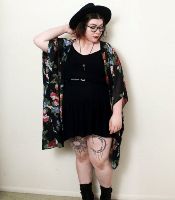 chica curvy usando lentes, sombrero negro de ala grande, vestido mini negro con cinturón negro, botines negros y kimono negro con flores; outfit estilo witchy