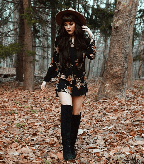 chica de cabello largo castaño usando un sombrero de ala grande café, vestido mini negro con flores rojas, botas largas de piso negras; outfit estilo witchy