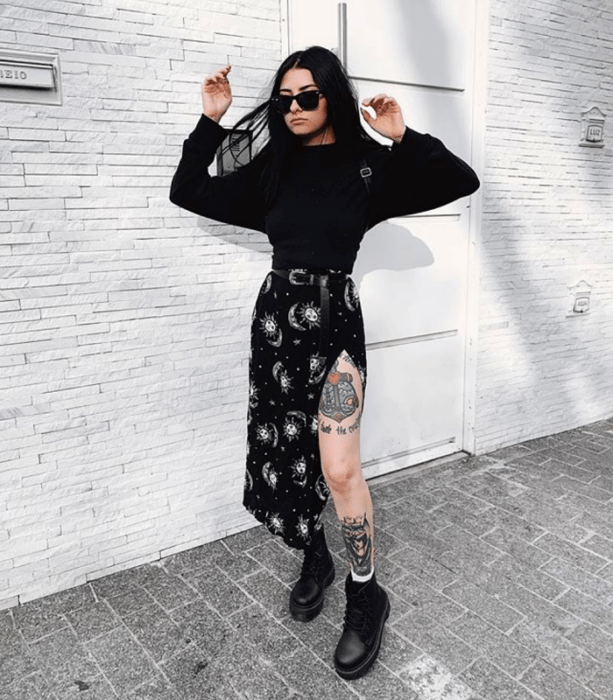 chica de cabello negro largo usando lentes de sol, blusa de manga larga negra, falda midi negra con estrellas y soles, botines negros de cuero