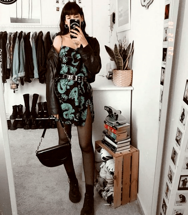 chica de cabello negro usando un vestido negro con dragones verdes, cinturón de estoperoles, medias negras, botines negros de cuero, bolso de mano negro y chaqueta de cuero