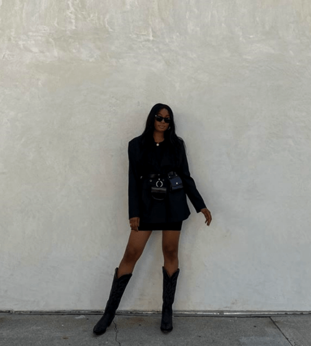 chica morena usando lentes de sol, saco oversized negro, falda negra, botas largas de cuero estilo vaquero y bolso de cuero negro a la cintura