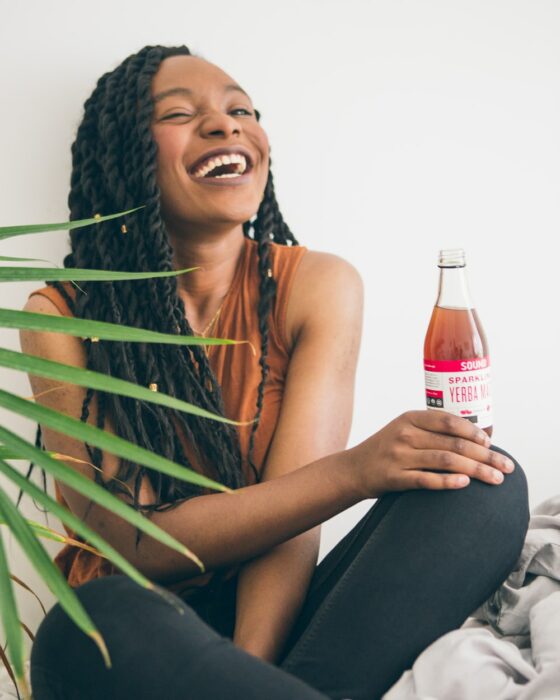 Chica de tez negra, con rastas sentada tomando un refresco y riéndo