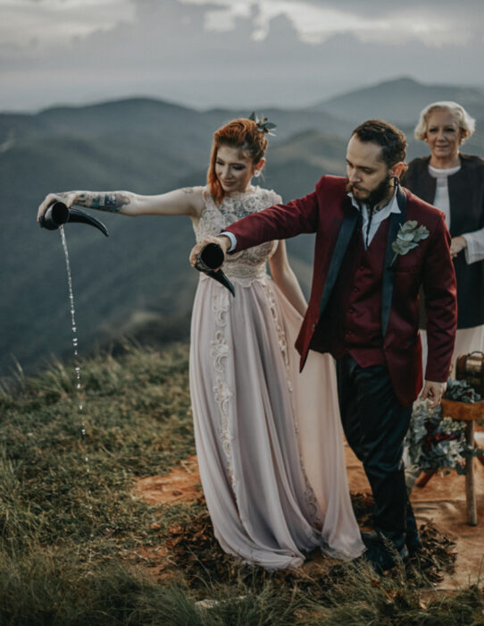 Ideas para una boda vikinga; novios con vasos de cuernos, vestido de encaje y saco rojo vino