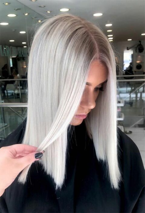 Chica con bata de estética negra mirando hacia abajo con melena lisa con raya en medio con tono de cabello blanco 'icy blonde'