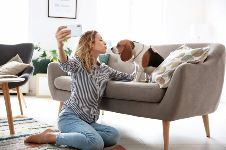 Los mejores celulares smartphone para tomar selfies bonitas; mujer rubia de blusa formal de rayas blancas y negras, pantalón de mezclilla azul claro, sentada en el suelo de la sala tomándose una fotografía con su perro beagle color blanco con manchas cafés y negras