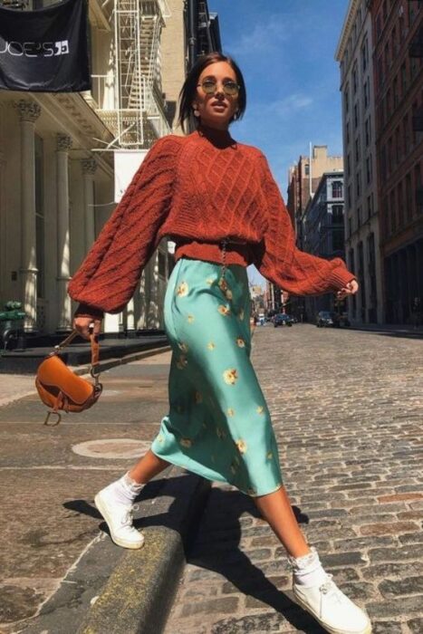 Chica de cabello corto caminando por la calle con falda de satín verde con flores, tenis blancos y suéter naranja