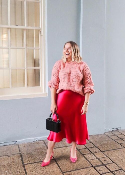 Chica plus size vrubia de cabello suelto ondulado vistiendo una falda de satín roja, suéter rosa y bolso de mano negro