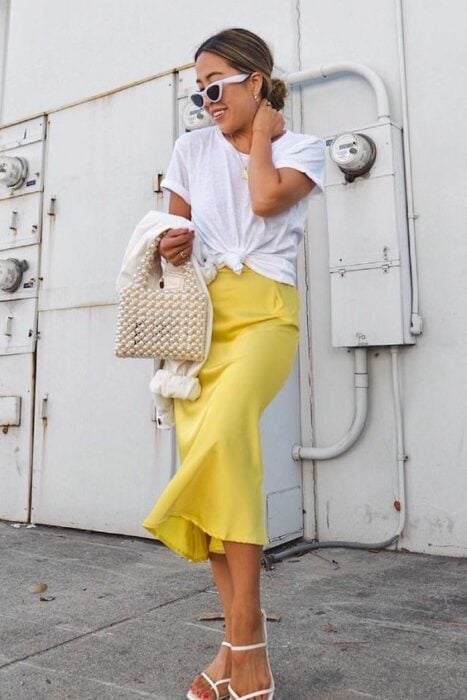 Chica rubia con una coleta pulida baja vistiendo blusa blanca, falda amarilla de satín y sandalias de tacon en tono nnude