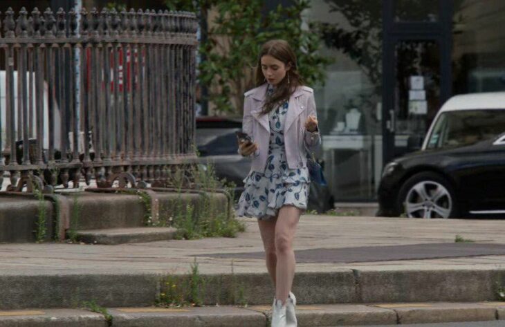 Lily Collins usando un atuendo de vestido floral con chaqueta de color morado con botas blancas en la serie Emily In Paris