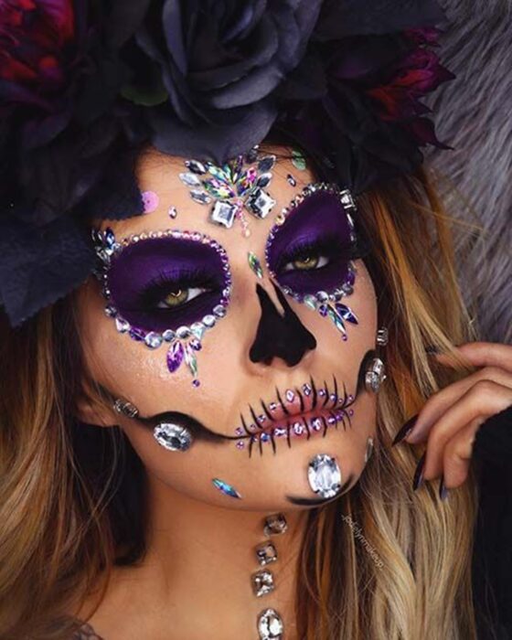 Chica con un maquillaje de catrina en colores morados con perlas y pedrería 