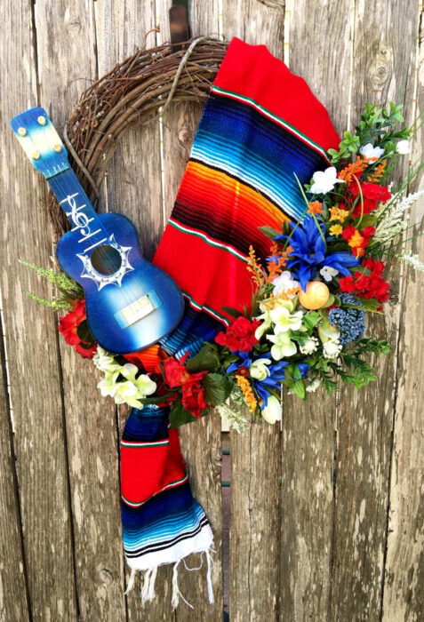 Decoración de Navidad con temática mexicana; corona para la puerta con ramas, reboso de colores rojo, azul y anaranjado, guitarra pequeña y flores de plástico